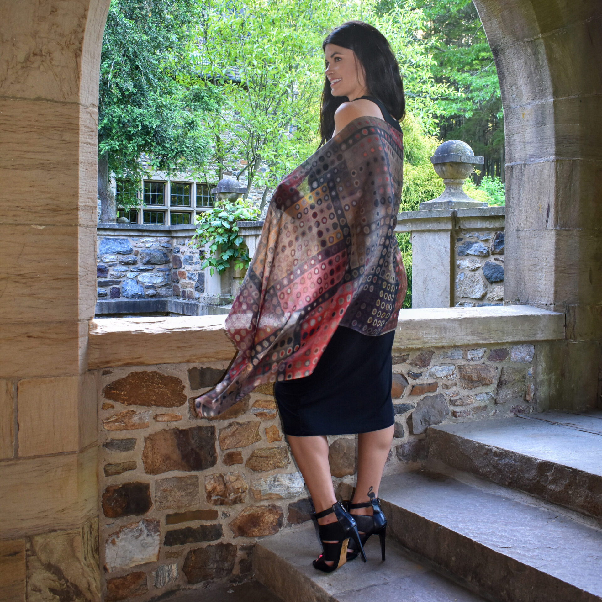 Woman in black dress and patterned shawl stands on stone steps by an old stone wall.